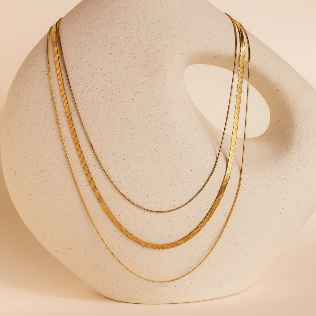 The Triple Layered Chain Necklace, featuring gold herringbone and snake chains, is elegantly draped over a modern abstract cream-colored ceramic sculpture with a handle.