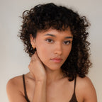 A woman with curly hair in a brown top gazes at the camera, her hand resting on her neck adorned with Dainty Opal Earrings. The scene is set against a neutral background.