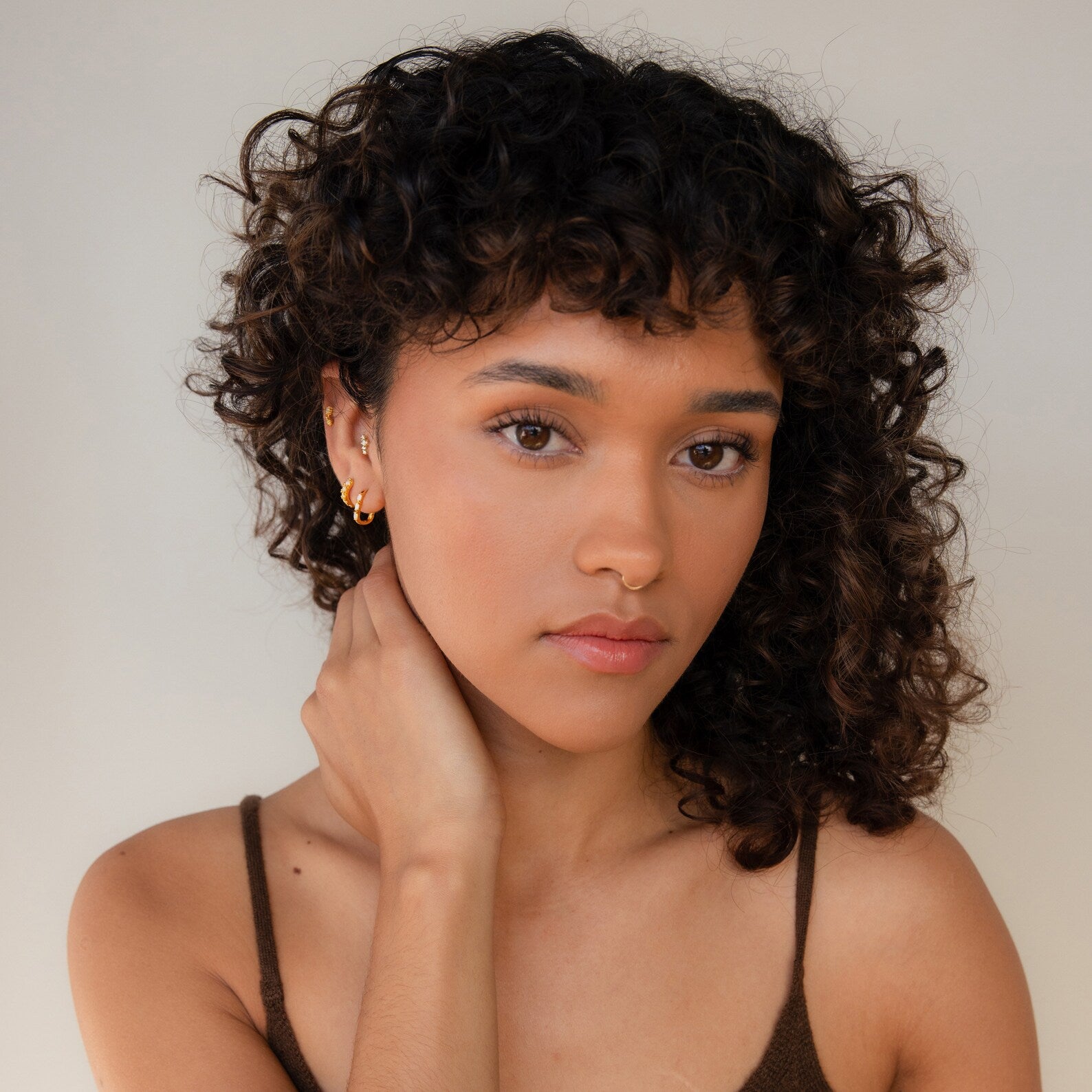 A woman with curly hair in a brown top gazes at the camera, her hand resting on her neck adorned with Dainty Opal Earrings. The scene is set against a neutral background.