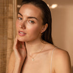 Woman with wet hair wears the Pearl Hummingbird Necklace in 18K Gold and gold earrings, touching her face in soft natural light.