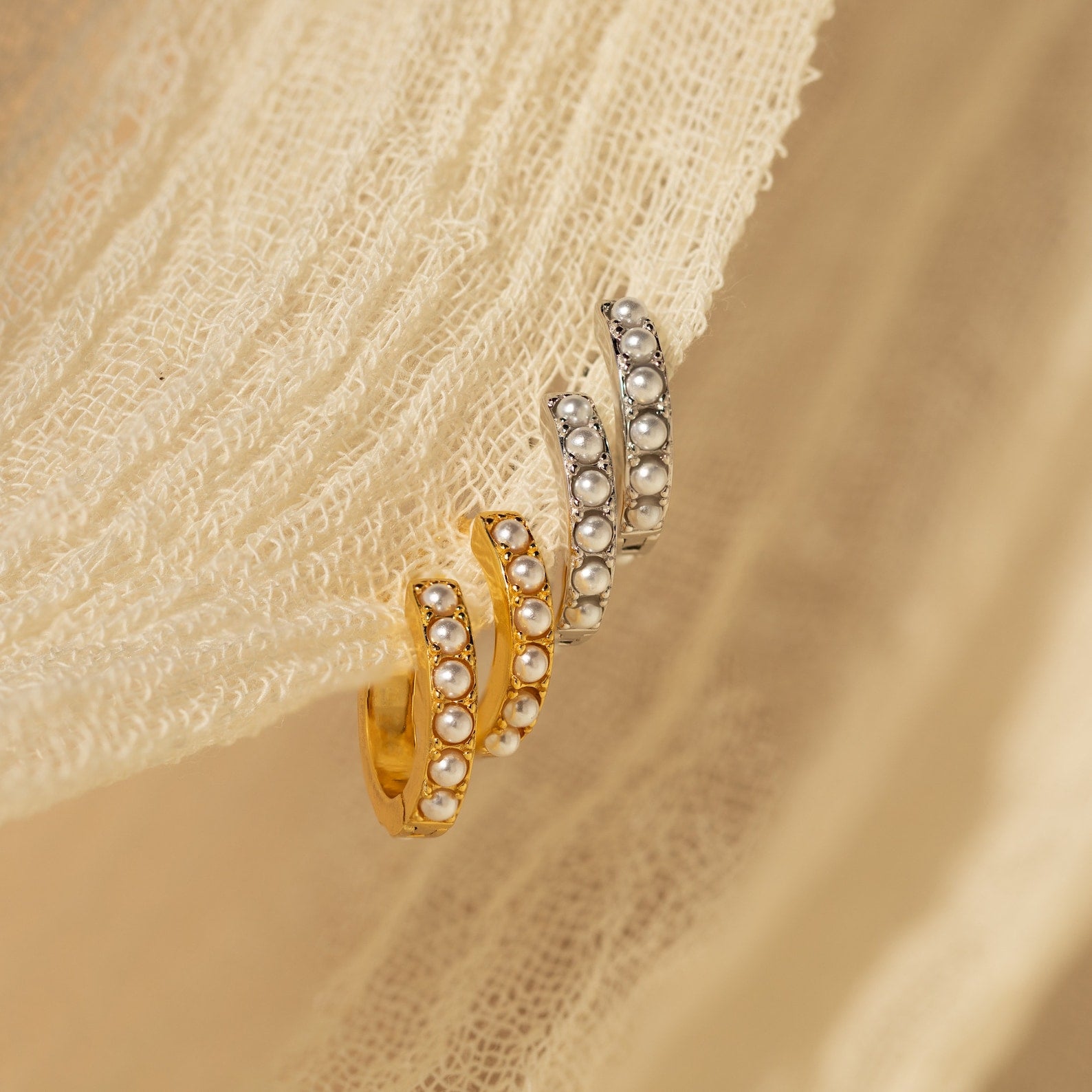 Two pairs of pearl-accented huggie earrings in gold and silver tones displayed side by side on beige fabric.

