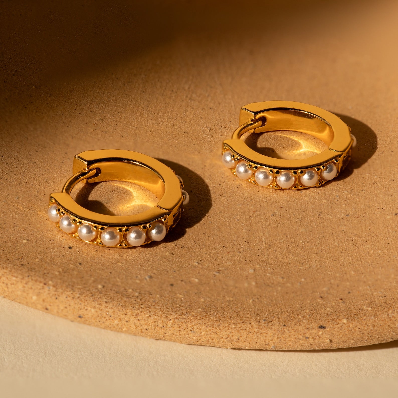Gold pearl huggie earrings resting on a warm-toned ceramic plate under direct sunlight.