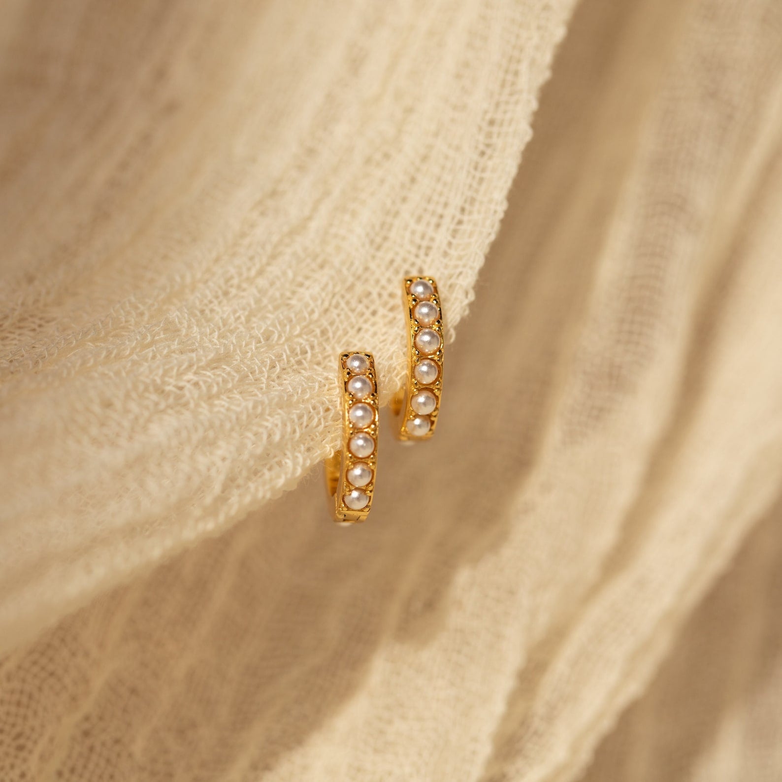A close-up of a pair of gold huggie earrings delicately draped over a soft, ivory textured fabric. The earrings feature a refined, symmetrical row of luminous miniature pearl beads set in a beaded, gold-finished channel. 