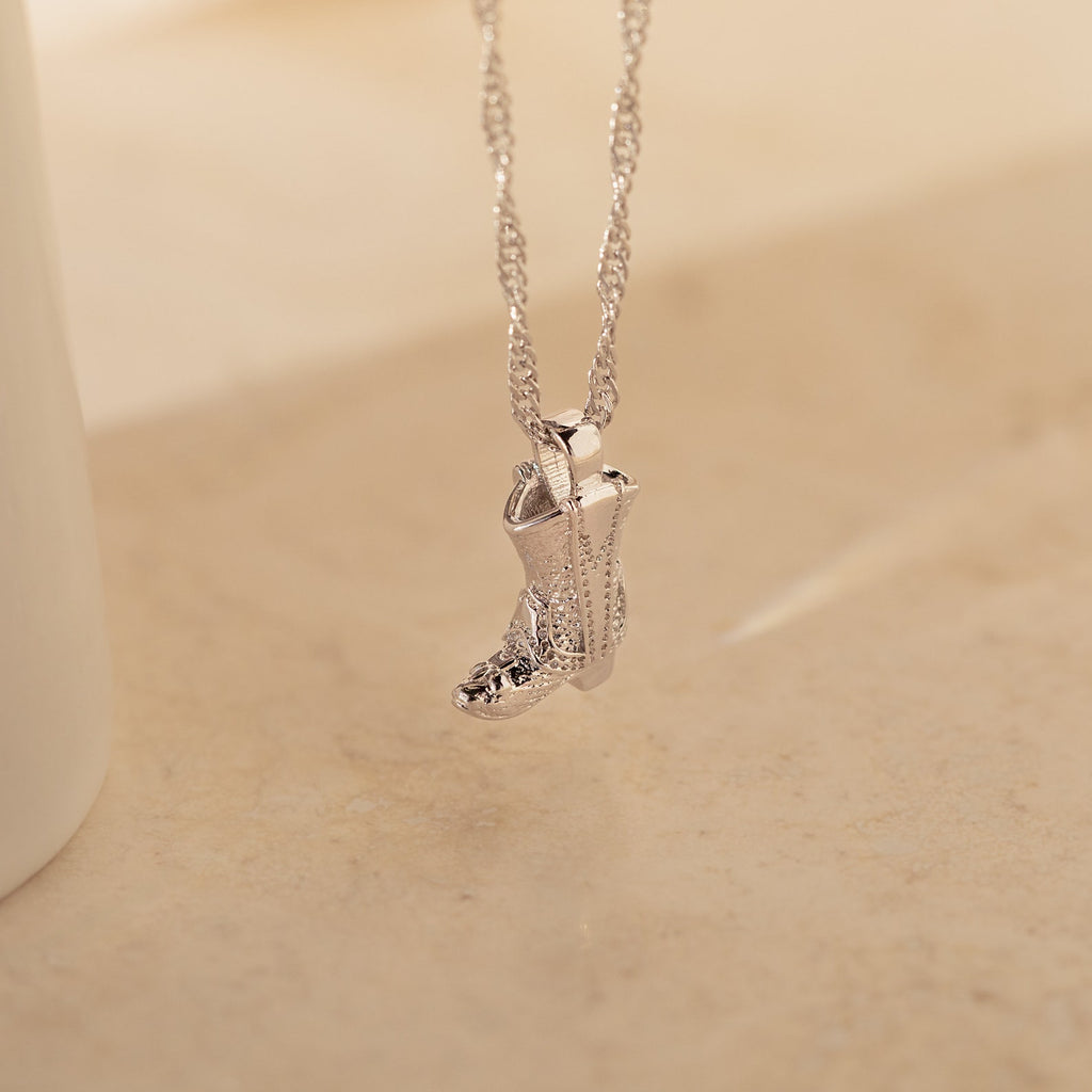The Cowboy Boot Necklace features a silver, engraved boot pendant that hangs elegantly against a beige background, showcasing timeless western charm.