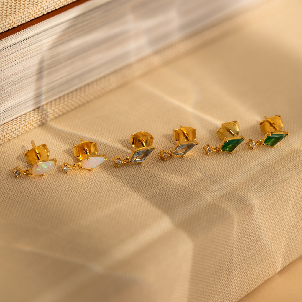 Three pairs of Art Deco Drop Studs—featuring opal, CZ diamond, and green stones—are displayed on a beige textured surface.