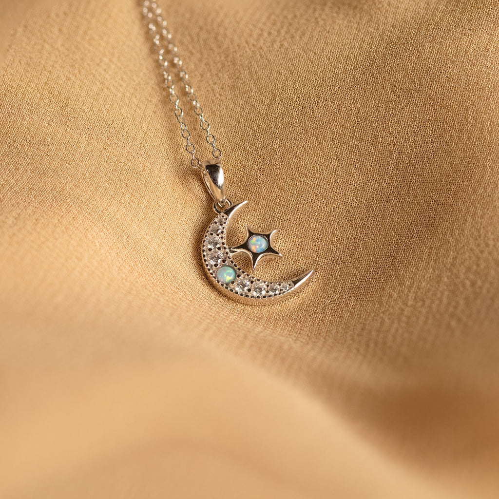 The Opal Moon & Star Necklace showcases a gold crescent moon and star pendant with opal details, elegantly displayed on a soft beige fabric background.