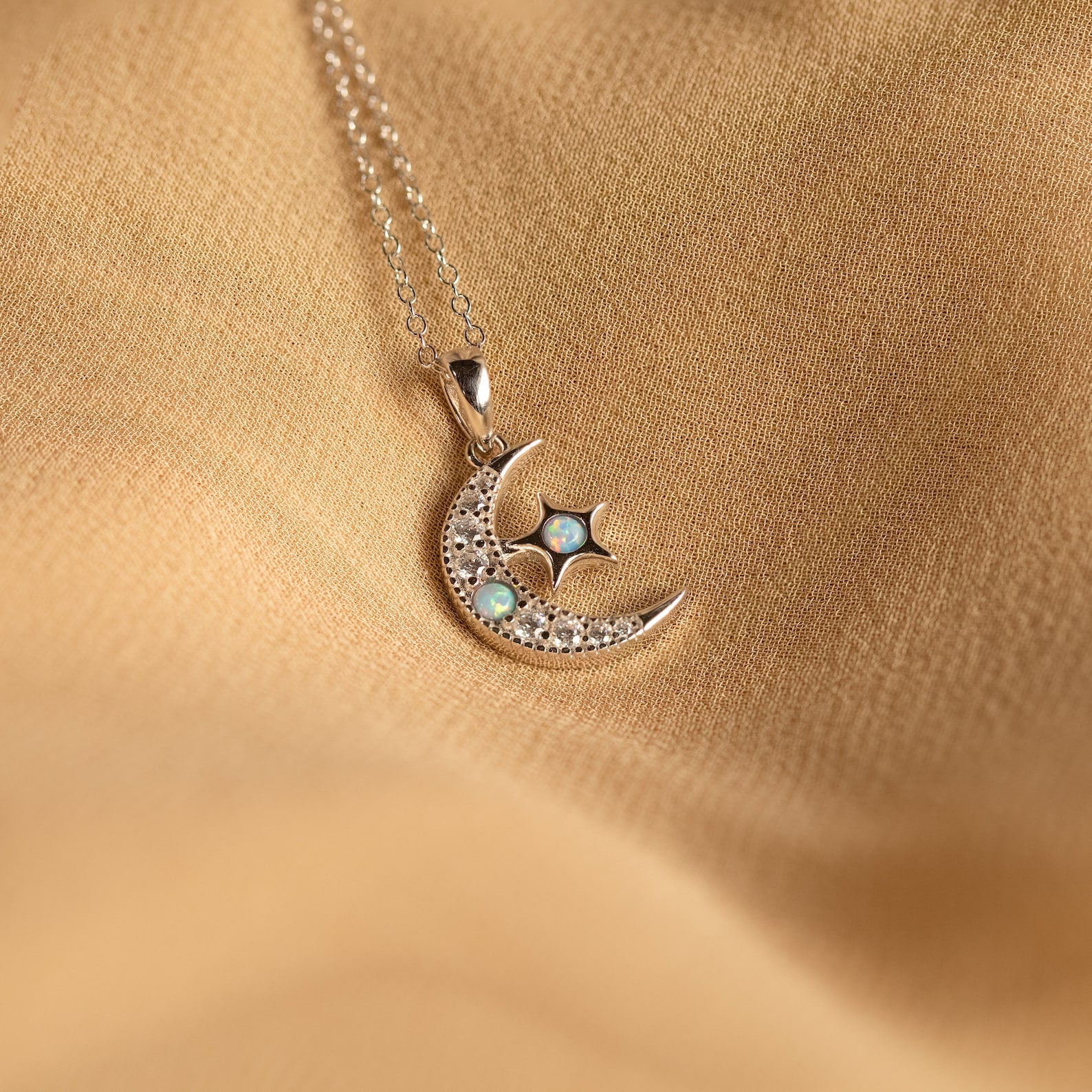 The Opal Moon & Star Necklace showcases a gold crescent moon and star pendant with opal details, elegantly displayed on a soft beige fabric background.