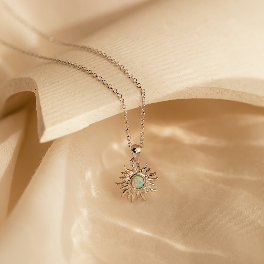 The Opal Sun Necklace features a silver sun-shaped pendant with a striking blue-green stone, beautifully presented on cream fabric—an elegant and thoughtful jewelry gift.