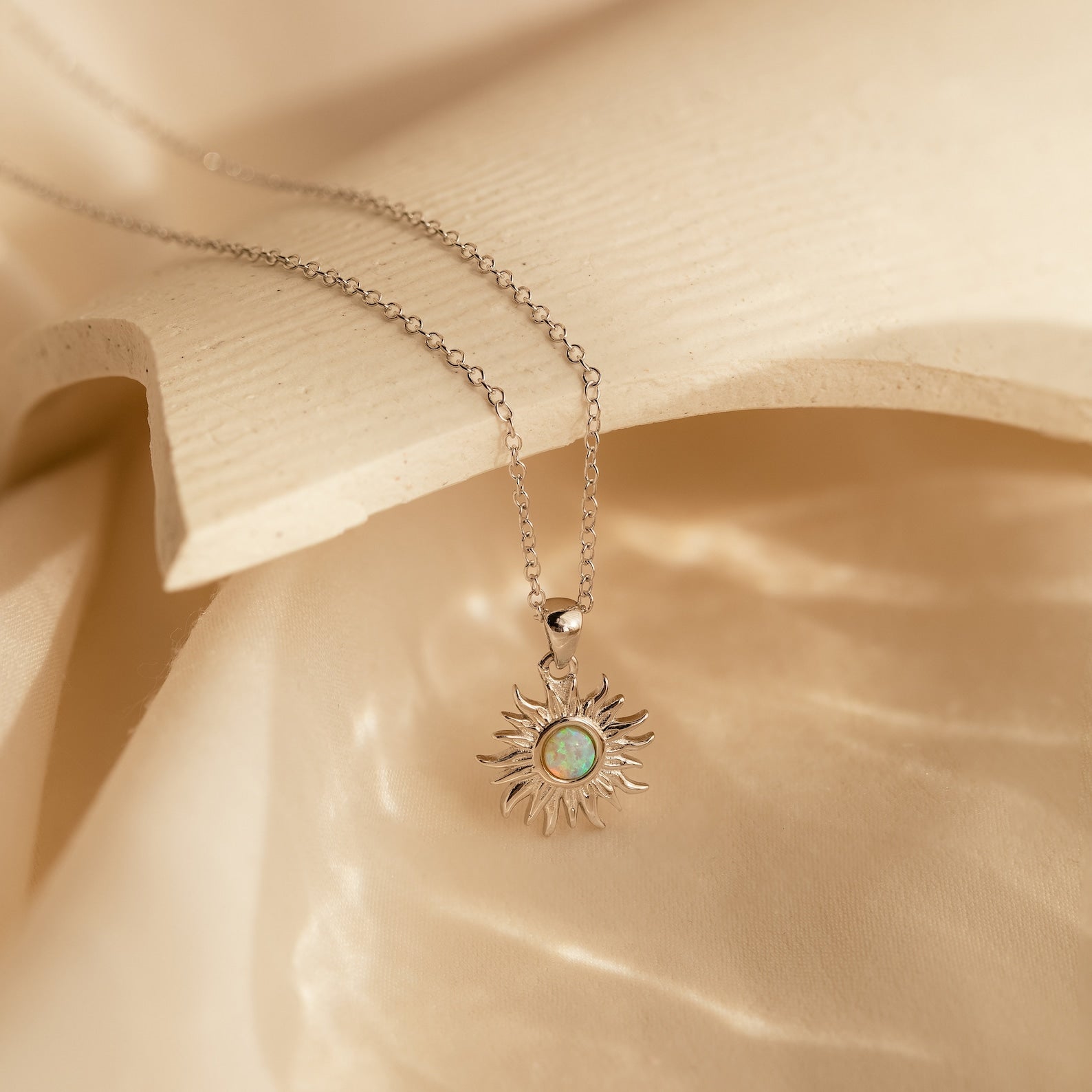 The Opal Sun Necklace features a silver sun-shaped pendant with a striking blue-green stone, beautifully presented on cream fabric—an elegant and thoughtful jewelry gift.