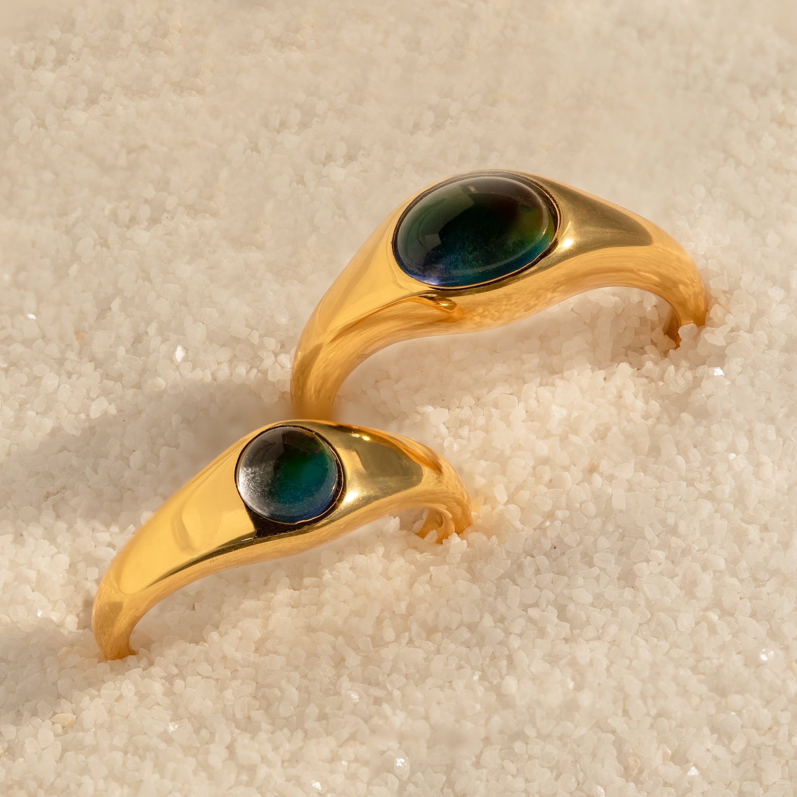 The Signet Mood Rings Set features two gold rings with oval dark stones that change color, displayed on white sand—a nostalgic addition to any jewelry collection.