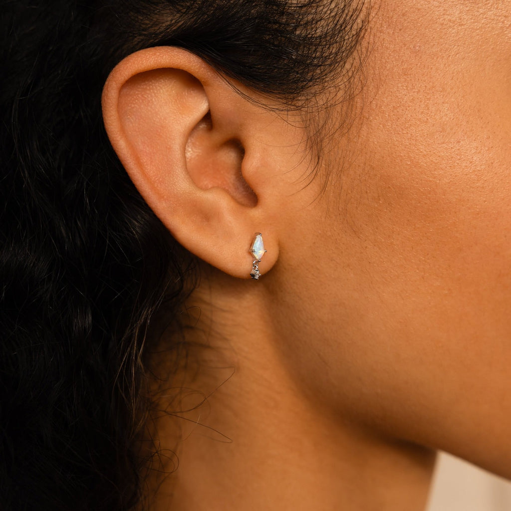 Close-up of a person’s ear wearing the Opal Kite Drop Earrings featuring a small, silver setting with a light blue stone and tiny CZ diamond accents in a subtle geometric design.
