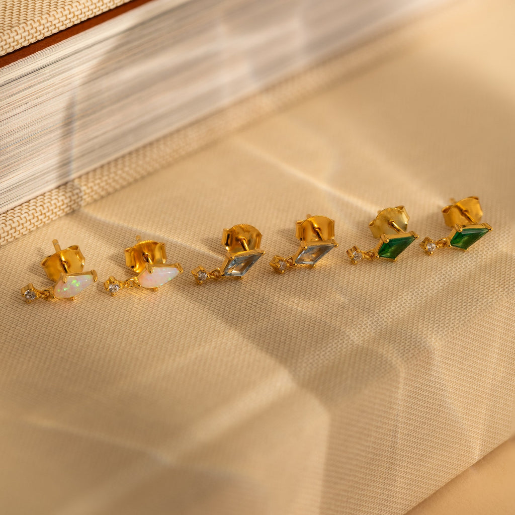 The Opal Kite Drop Earrings—gold with opal and geometric design—are displayed on beige fabric.