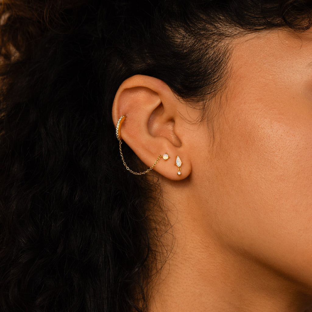 Close-up of an ear with three gold earrings, one being the Opal Kite Drop Earrings featuring a delicate chain and accented with a CZ Diamond in a bold geometric shape.