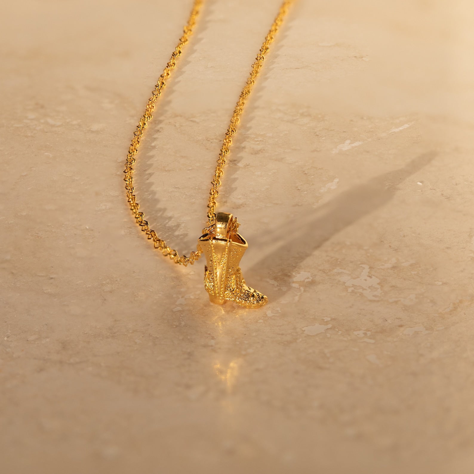 Cowboy Boot Necklace with an engraved gold boot pendant, showcased on a smooth beige surface for a stylish western accent.