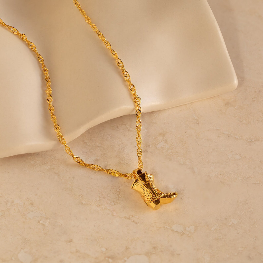 Cowboy Boot Necklace features a gold chain and engraved boot pendant, shown on beige marble. Ideal for bringing western style to any outfit.