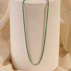 The Emerald Tennis Choker Necklace is elegantly displayed on a white cylindrical stand with a beige fabric background—ideal for layering or wearing solo as a stunning statement piece.