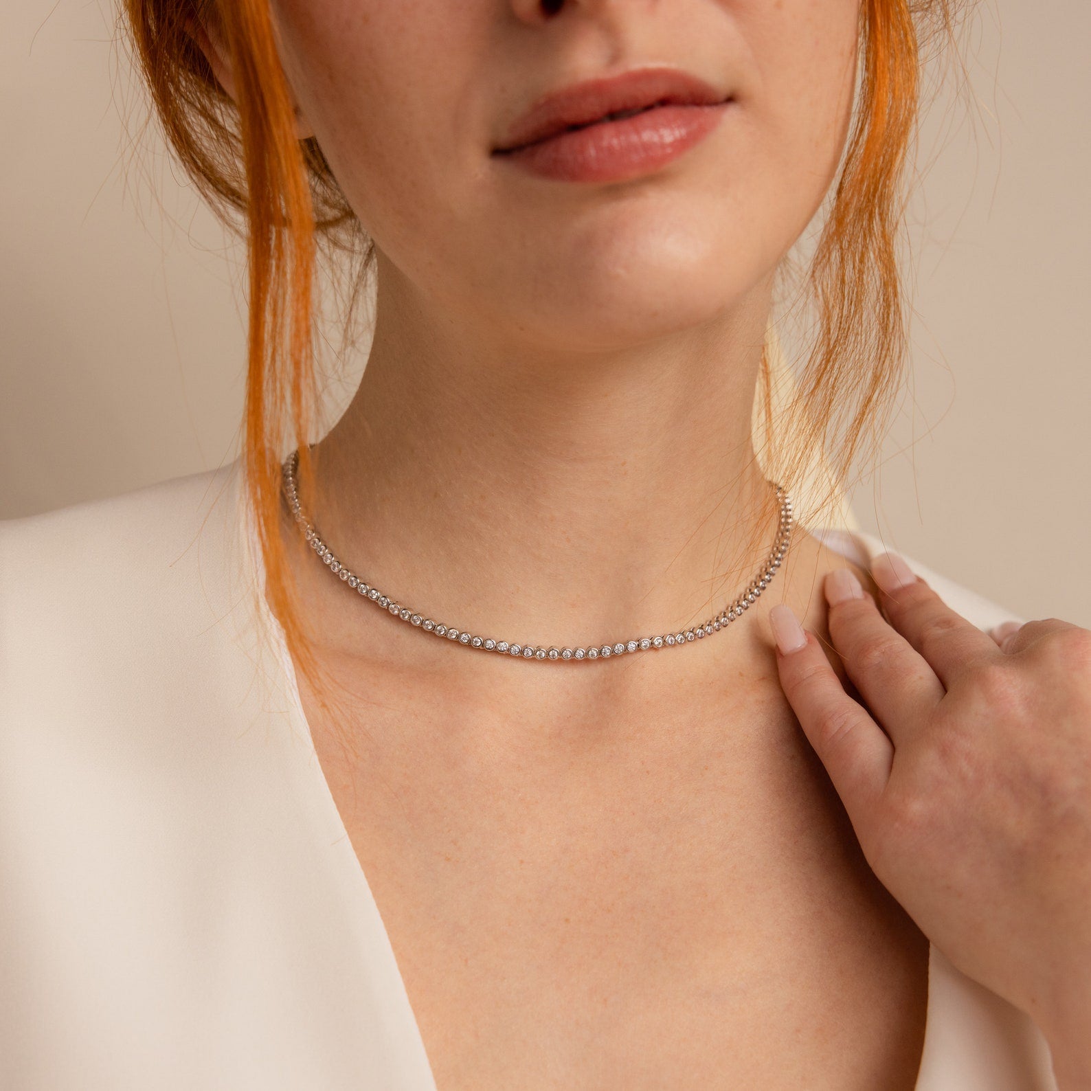 A woman in a white top wears the Diamond Bezel Tennis Necklace & Bracelet Set in White Gold, gently touching her collarbone.