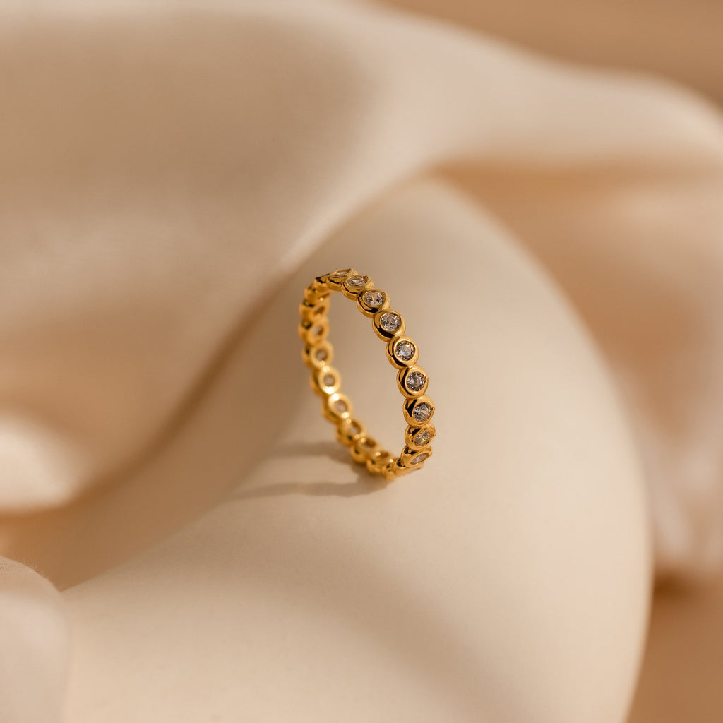 The Diamond Bezel Tennis Ring features small round CZ gemstones in a continuous setting, presented on a soft beige fabric background.