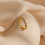 The Diamond Bezel Tennis Ring features small round CZ gemstones in a continuous setting, presented on a soft beige fabric background.