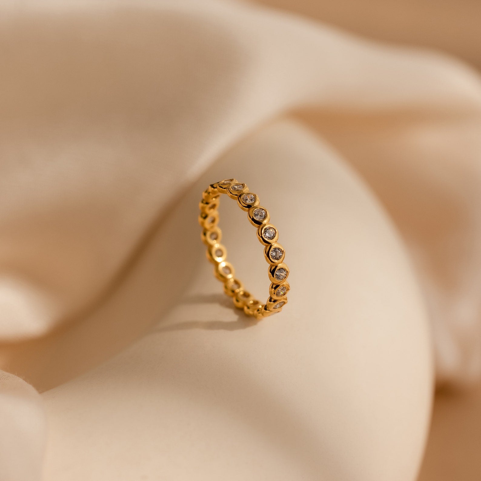 The Diamond Bezel Tennis Ring features small round CZ gemstones in a continuous setting, presented on a soft beige fabric background.