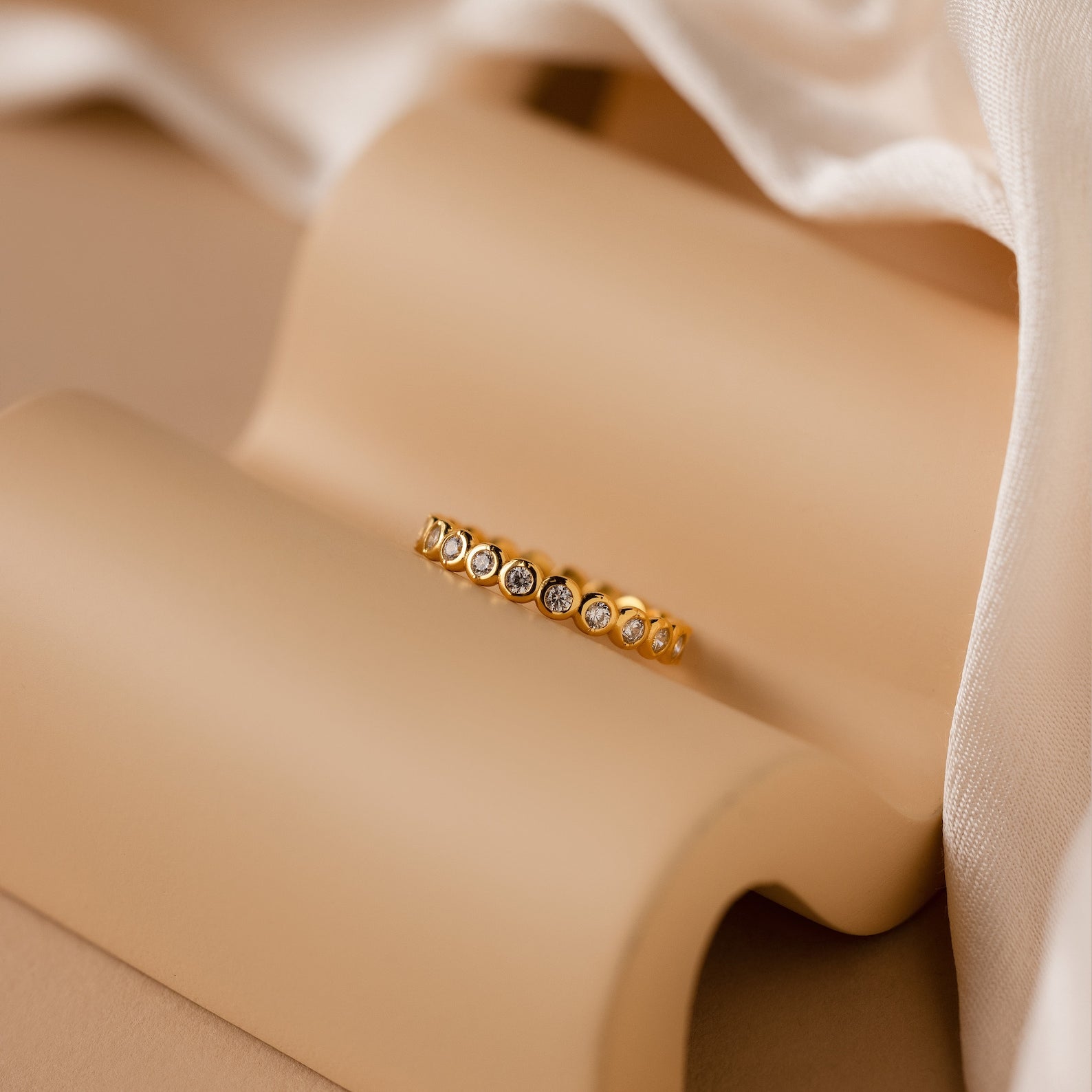 Diamond Bezel Tennis Ring featuring round, clear CZ gemstones displayed on a beige curved surface with soft fabric in the background.