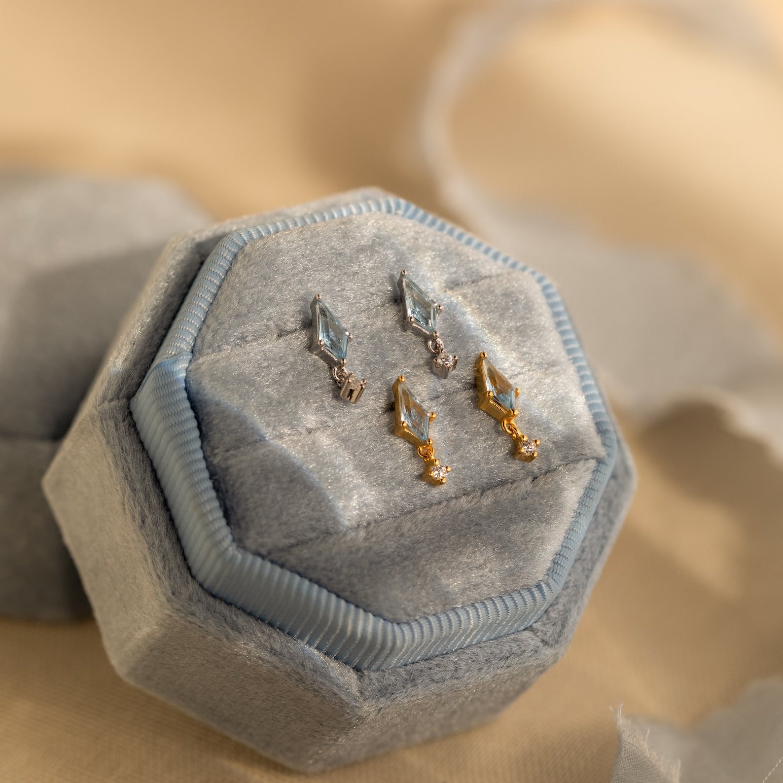 Two pairs of geometric gemstone earrings, including Art Deco Drop Studs and CZ Diamond designs in silver and gold, displayed on a light blue velvet ring box.