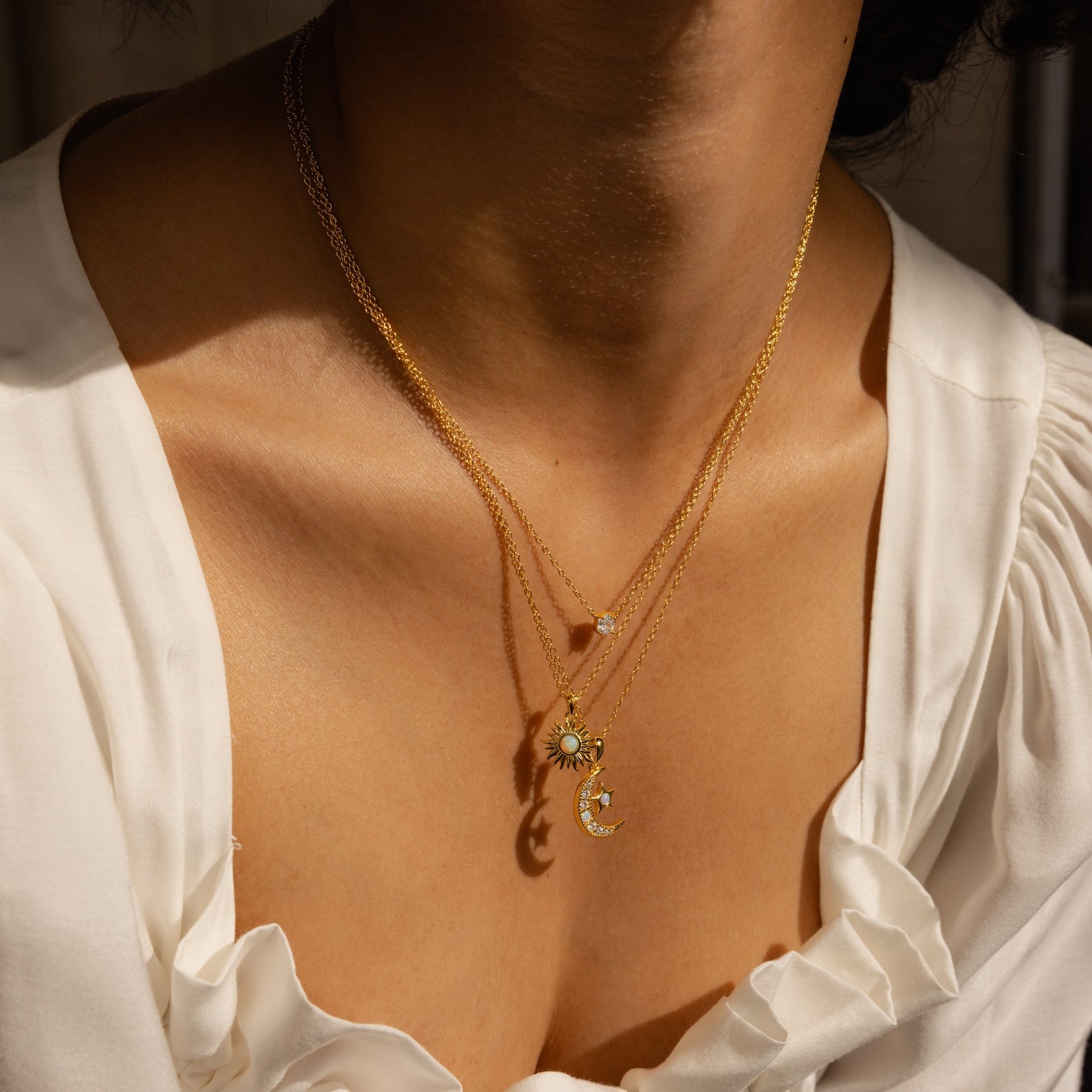 Close-up of a woman wearing the Celestial Pendant Necklace Set—layered gold necklaces with sun, moon, and crystal charms—styled over a white blouse; an elegant gift for her.