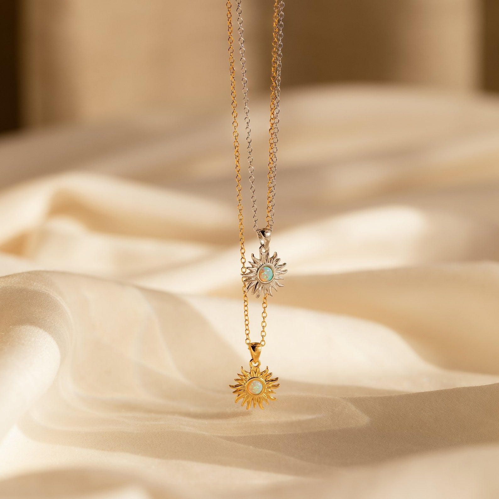 The Opal Sun Necklace features two sun pendants—one gold, one silver—with blue centers on delicate chains, displayed on soft beige fabric. This dainty piece makes a stunning jewelry gift.