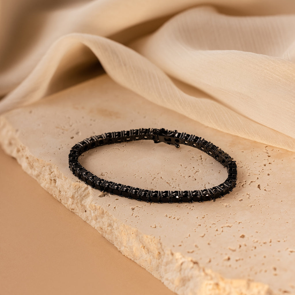 The Onyx Tennis Bracelet is showcased on a textured stone surface, with a beige fabric draped in the background.