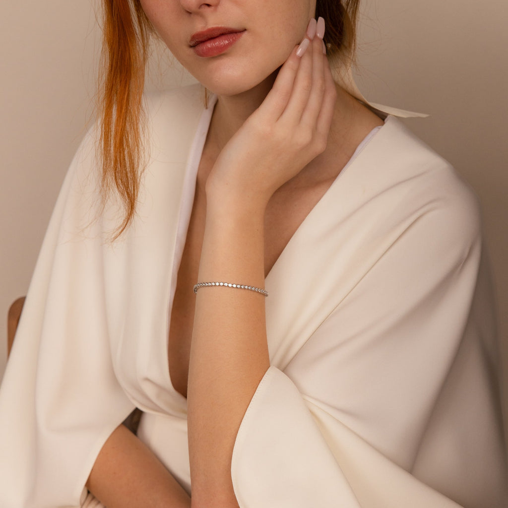 Woman in a white dress wearing the Diamond Bezel Tennis Necklace & Bracelet Set in White Gold, hand gently touching her chin against a neutral background—an elegant choice for an anniversary gift.
