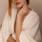 Woman in a white dress wearing the Diamond Bezel Tennis Necklace & Bracelet Set in White Gold, hand gently touching her chin against a neutral background—an elegant choice for an anniversary gift.