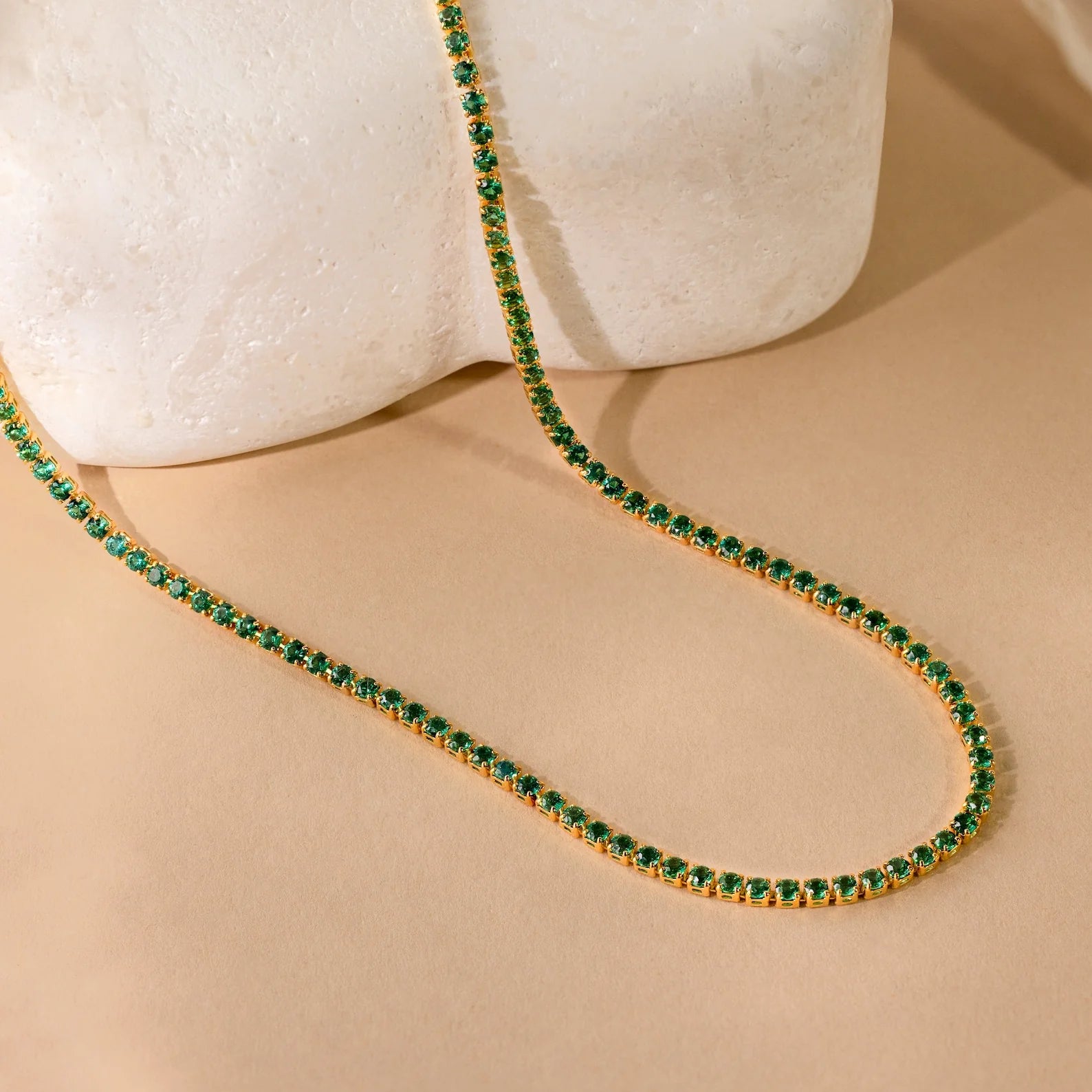 A Tennis Necklace Set with green gemstones is displayed on a light stone against a beige background, ideal for layering jewelry.