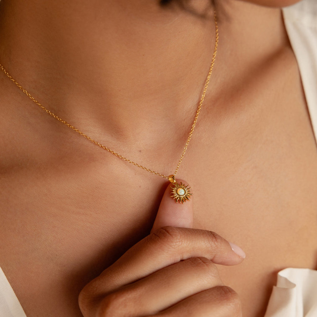 A person holds the Celestial Pendant Necklace Set featuring a gold sunburst design with a white opal and diamond center on their chest—the perfect gift for her.