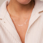 A woman wears the Summer Tiny Multiple Name Necklace, personalized with "Sierra Kendall Bailey" in elegant cursive letters, paired with a cream sweater.