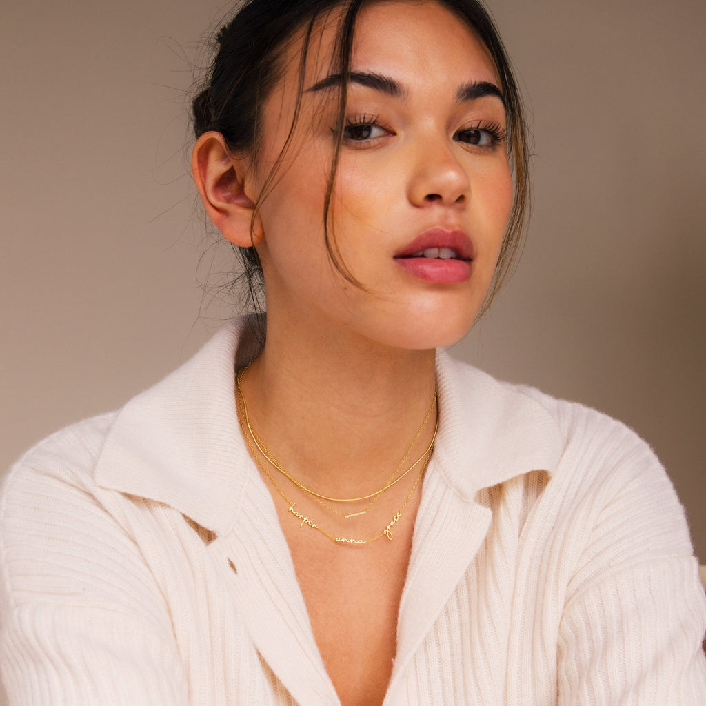 Woman in a cream ribbed top wears layered gold necklaces, including the Summer Tiny Multiple Name Necklace, and looks confidently at the camera.