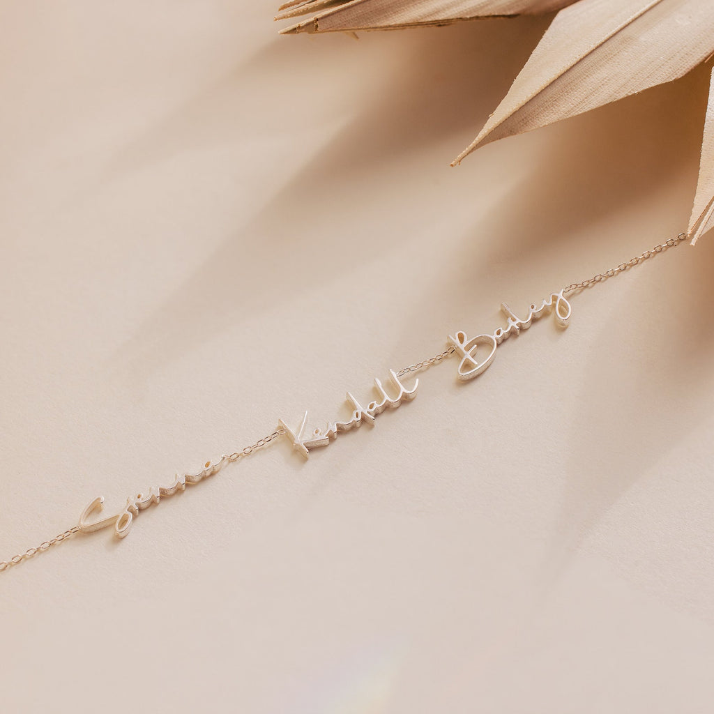 The Summer Tiny Multiple Name Necklace features elegant cursive letters and is styled on a neutral beige background with dried leaves—an ideal piece of custom jewelry for those who love personalized necklaces.