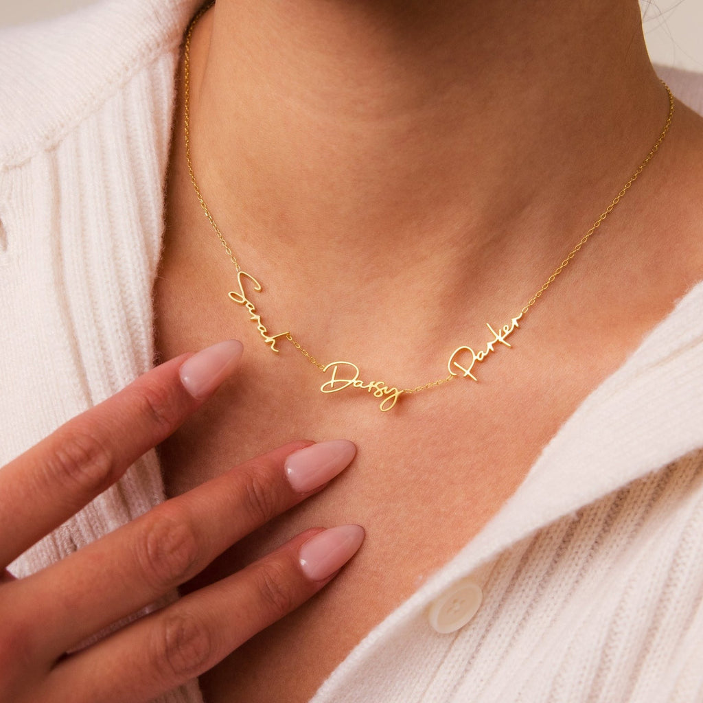 A woman wears the Summer Tiny Multiple Name Necklace in gold, personalized with the names Sarah, Daisy, and Parker in elegant cursive, resting gently against her collar.