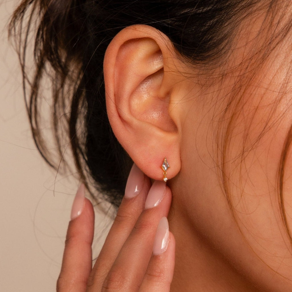 Close-up of a woman's ear wearing Aquamarine Kite Drop Earrings, featuring a small gold geometric design, as her manicured hand touches her earlobe.