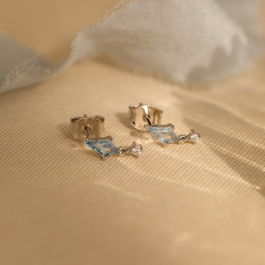 Aquamarine Kite Drop Earrings with small dangling blue gemstones in a geometric design, accented by tiny CZ diamonds, showcased on a beige fabric background.