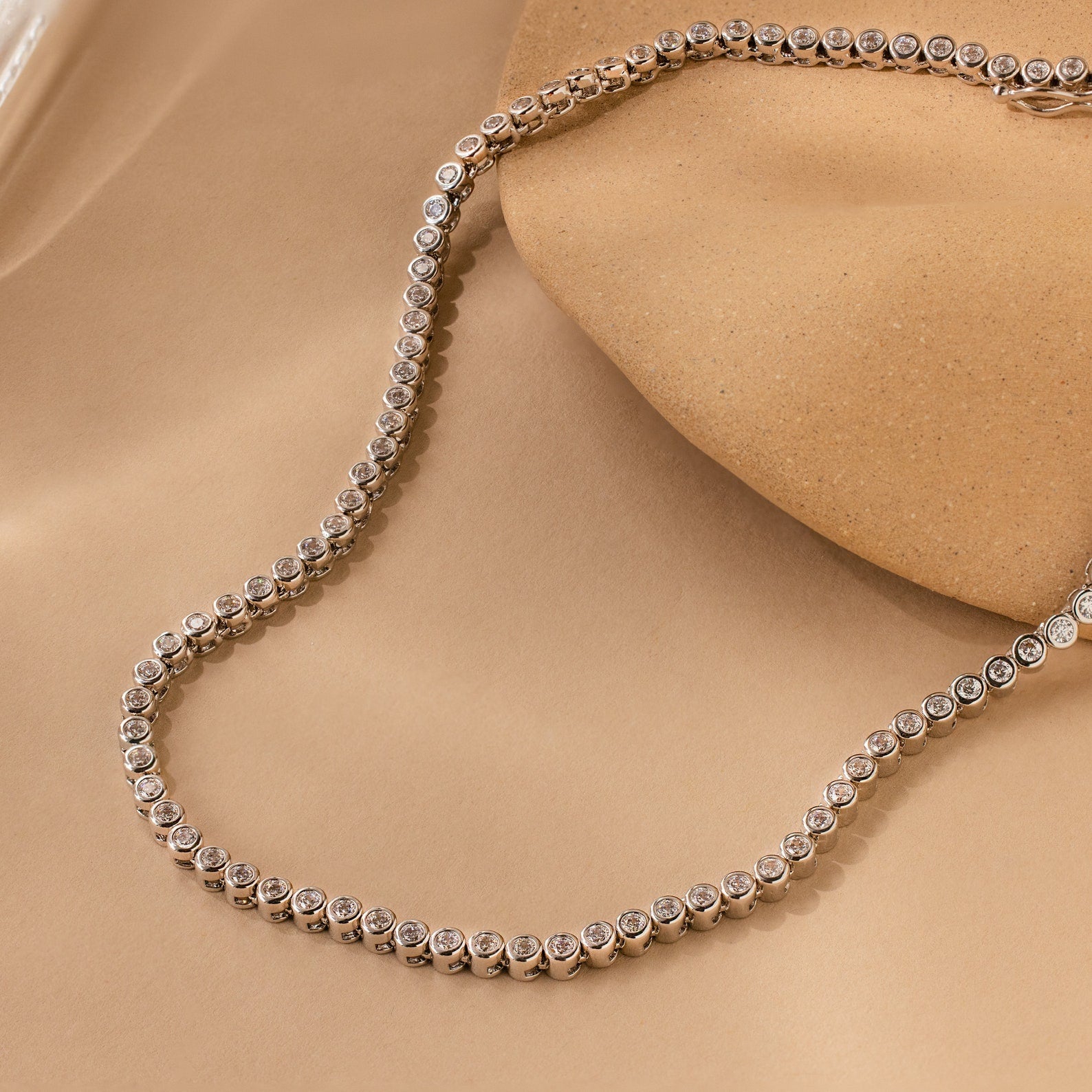 A Diamond Bezel Tennis Necklace & Bracelet Set in white gold, featuring round diamonds and displayed on a beige textured background—an elegant anniversary gift.