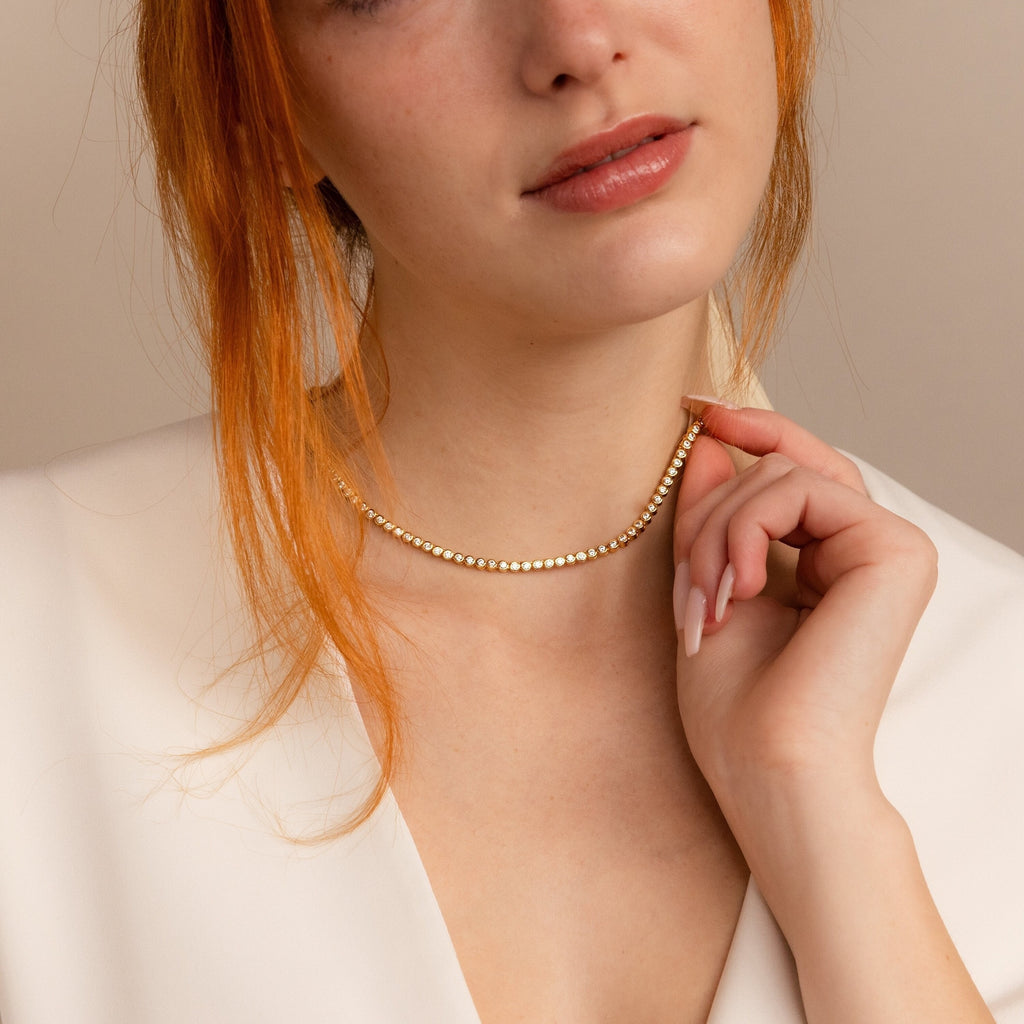 A woman in a white top holds the Diamond Bezel Tennis Necklace & Bracelet Set in 18K Gold, showcasing its elegant design under soft lighting with a neutral background.