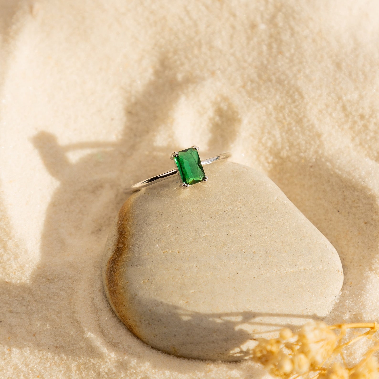 The Harley Emerald Ring, a vintage-style solitaire featuring a rectangular green gemstone, rests atop a smooth stone in the sand.