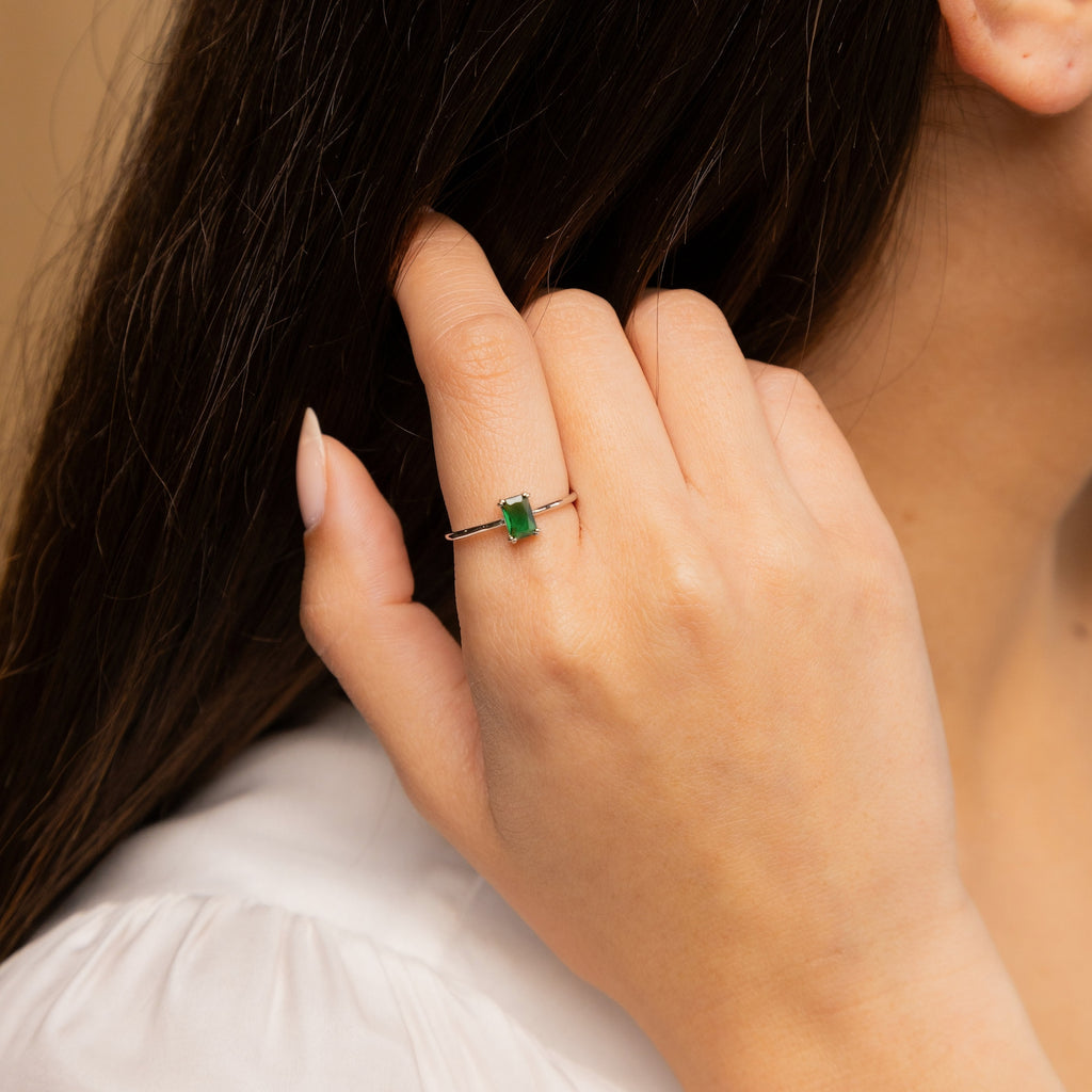 A woman wears the Harley Emerald Ring - Silver, featuring a rectangular green gemstone on her finger near her face—a beautiful nod to vintage-inspired style.