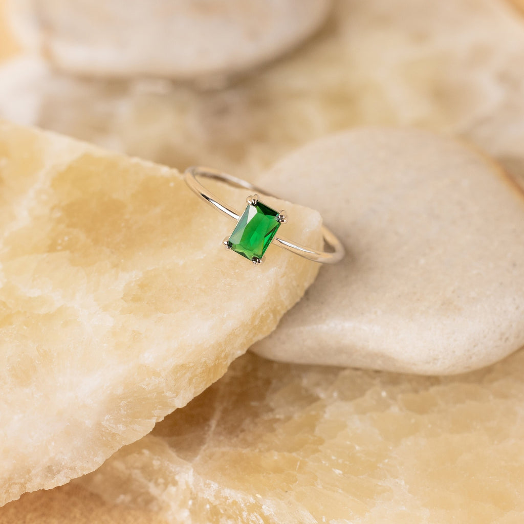 The Harley Emerald Ring - Silver features a rectangular green gemstone in an Art Deco-inspired design, reminiscent of vintage jewelry, and is beautifully displayed on beige and cream-colored stones.