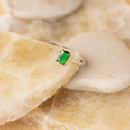 The Harley Emerald Ring - Silver features a rectangular green gemstone in an Art Deco-inspired design, reminiscent of vintage jewelry, and is beautifully displayed on beige and cream-colored stones.