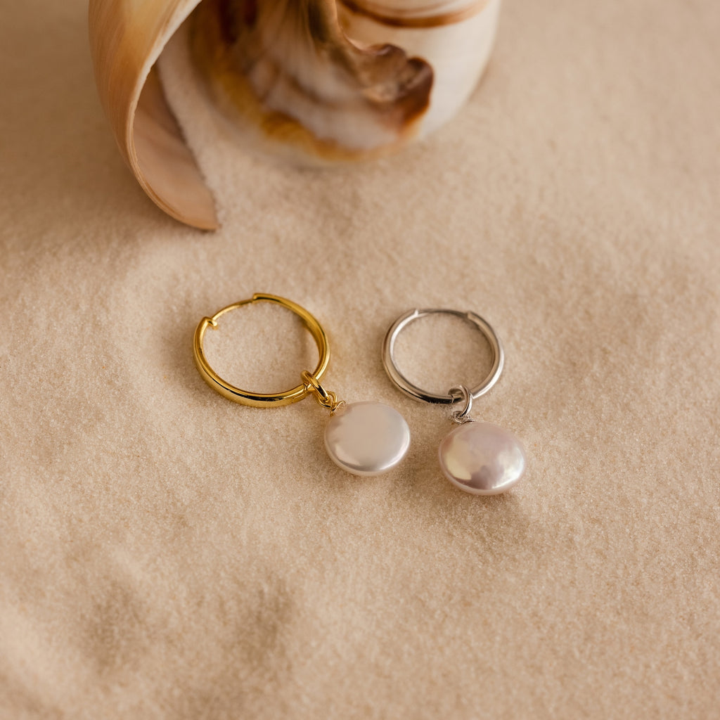 The Round Pearl Charm hoop earrings in gold and silver rest on sand next to a seashell.