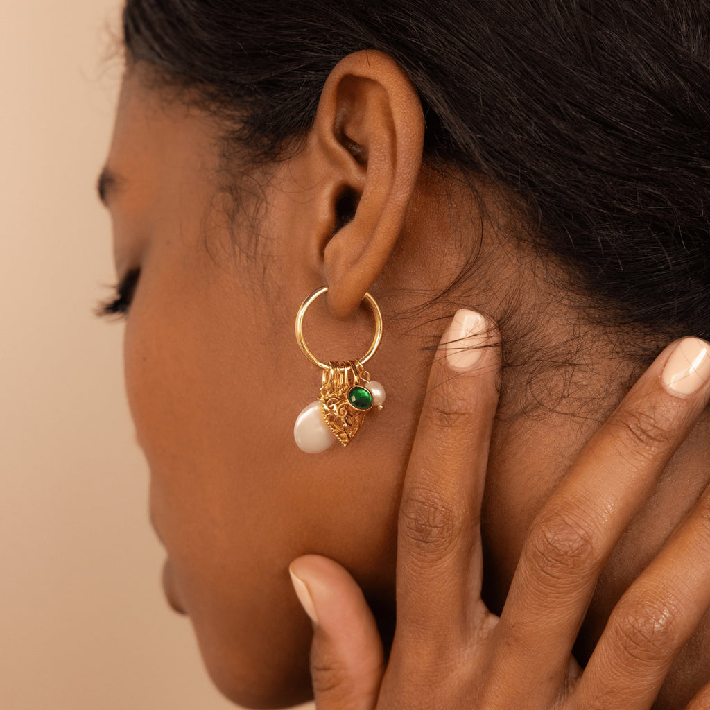 A woman touches her ear, wearing the Round Pearl Charm in 18K Gold—elegant gold hoop earrings adorned with a round pearl charm and green stone accents.