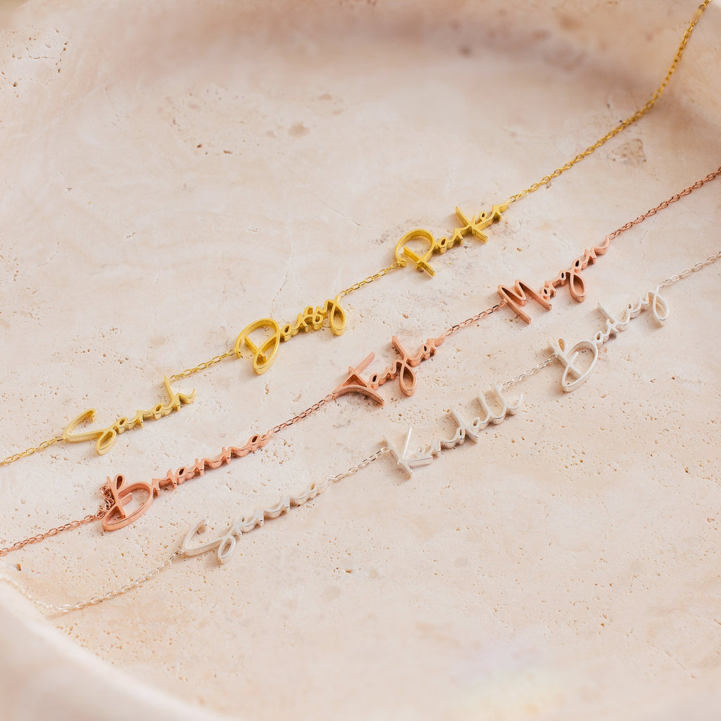 The Summer Tiny Multiple Name Necklace is shown in gold, rose gold, and silver on a light beige textured surface, ideal for anyone looking for elegant personalized jewelry.