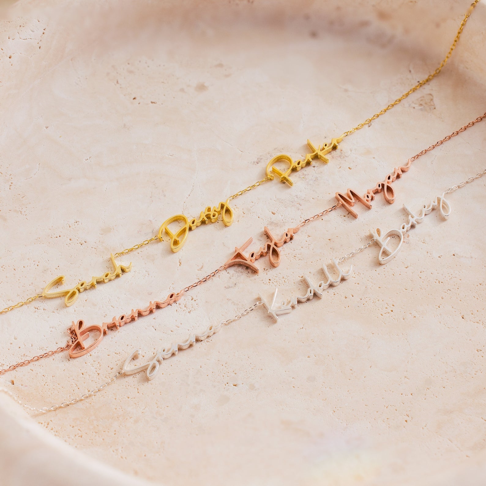 The Summer Tiny Multiple Name Necklace is shown in gold, rose gold, and silver on a light beige textured surface, ideal for anyone looking for elegant personalized jewelry.