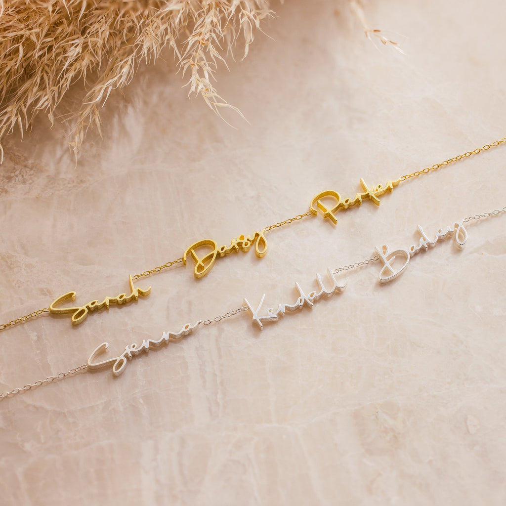 Two Summer Tiny Multiple Name Necklaces, one in gold and one in silver, rest on a marble surface with dried grass accents nearby.